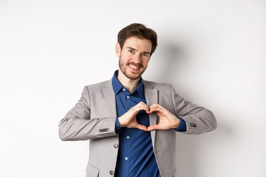 Romantic man in suit showing heart sign and smiling, love his girlfriend, standing on white background, making confession to lover.