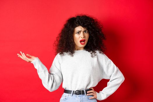 Annoyed and confused caucasian woman with curly hair, raising hand up and arguing, having argument, cant understand something, standing on red background.