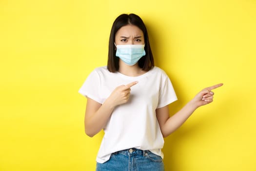 Covid-19, pandemic and social distancing concept. Disappointed asian girl in medical mask, frowning upset and pointing fingers right at logo, standing over yellow background.