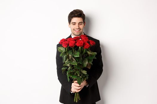 Image of handsome boyfriend in black suit, holding bouquet of red roses and smiling, being on a date, standing over white background.