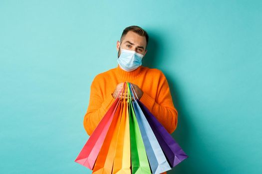 Covid-19, pandemic and lifestyle concept. Displeased and reluctant man in medical mask, holding shopping bags and complaining, standing over turquoise background.