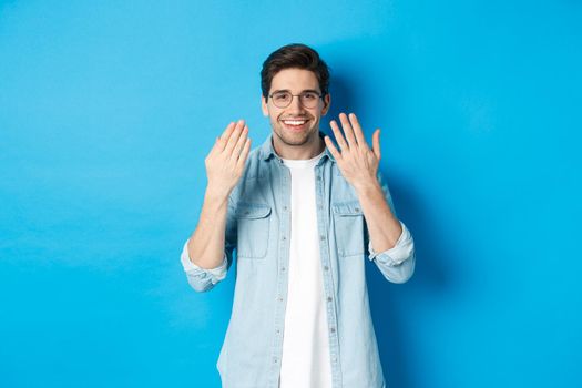Handsome young man in glasses showing manicure hands and smiling, taking care of nails, standing over blue background.