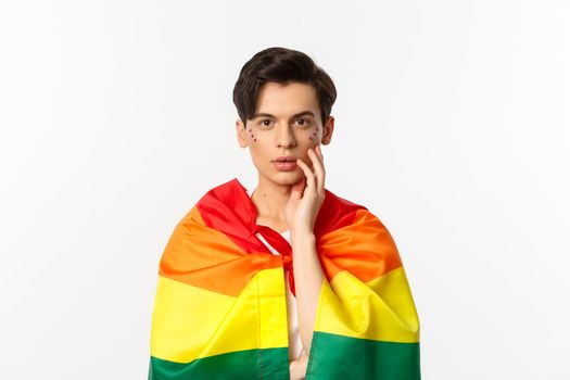 Beautiful young gay man wearing lgbt pride flag and glitter on face, touching cheek and gazing sensual at camera, white background.