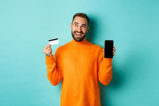 Online shopping. Thoughtful man shop in internet, showing mobile screen and credit card, looking at upper left corner and thinking, standing over light blue background.