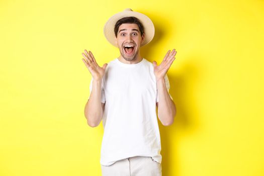 Concept of tourism and summer. Happy young man in straw hat looking amazed, reacting to surprise, standing over yellow background.