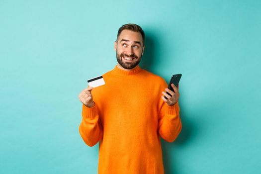 Online shopping. Handsome man thinking, holding smartphone with credit card, paying in internet store, standing over light blue background.