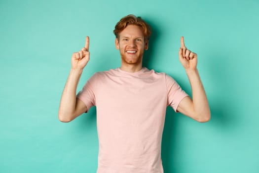 Handsome caucasian man with white teeth, smiling happy and pointing fingers up, showing promo offer, standing over mint background.