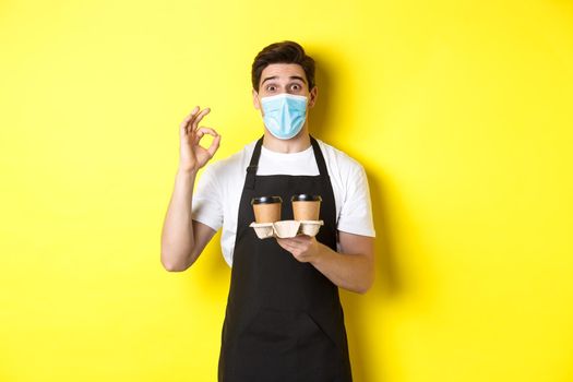 Concept of covid-19, cafe and social distancing. Barista in medical mask and black apron guarantee safety, holding takeaway cups of coffee and showing OK sign, yellow background.