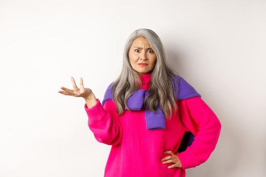 Angry and confused asian senior woman spread hand sideways and staring at camera puzzled, standing in pink sweater against white background.