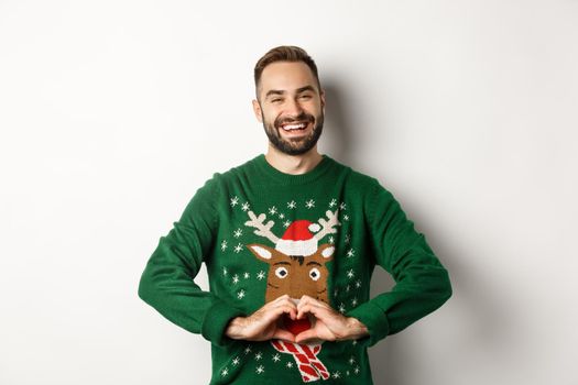 New Year, holidays and celebration. Happy bearded man in Christmas sweater showing heart sign, express love and care, standing over white background.