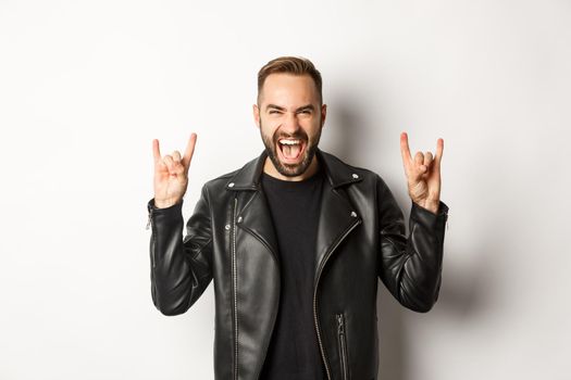 Cool adult man in black leather jacket, showing rock on gesture and tongue, enjoying music festival, standing over white background.