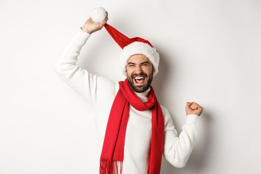 Winter holidays and New Year party concept. Happy man celebrating Christmas dancing in Santa hat and red scarf, standing against white background.