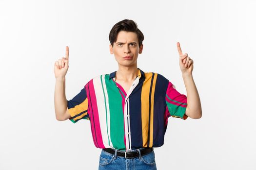 Skeptical and doubtful young gay man smirking, pointing and staring up at promo, hesitating, standing over white background.