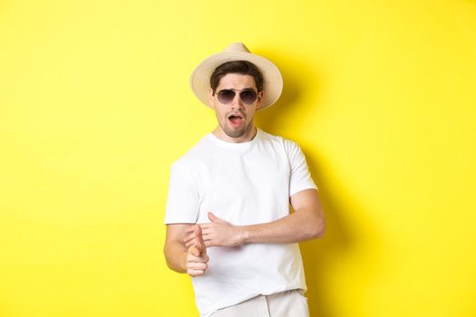 Handsome guy making finger gun shot and looking sassy, dressed for summer vacation in straw hat and sunglasses, standing against yellow background.