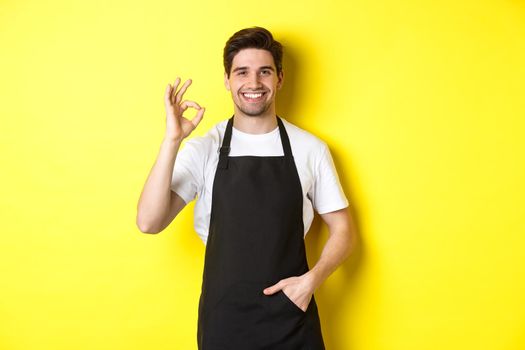 Young smiling barista in black apron showing okay sign, recommending coffee shop or restaurant, standing over yellow background.