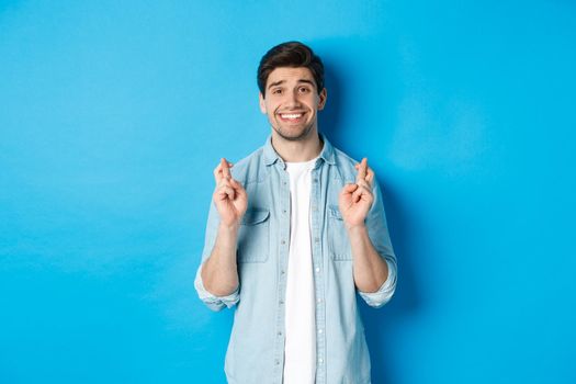 Handsome hopeful man making a wish, crossing fingers and smiling, waiting for results, standing against blue background.