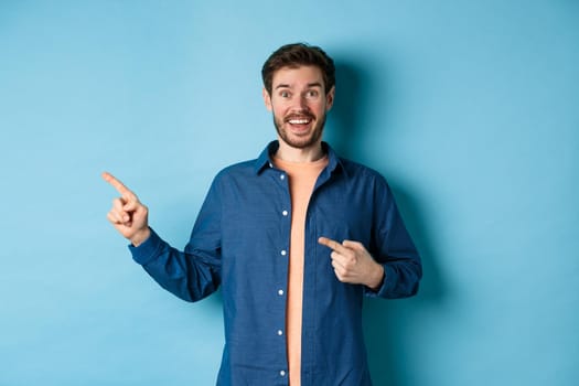 Excited and happy caucasian man smiling at camera, pointing fingers left at empty space, showing logo, standing on blue background. Copy space
