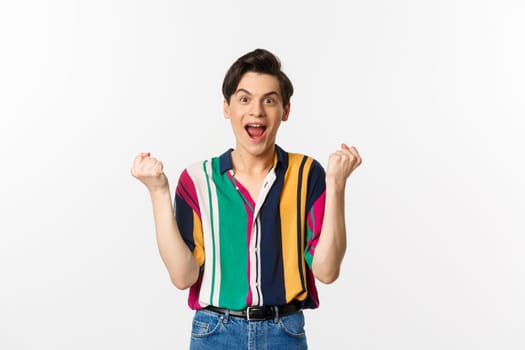 Image of successful young man achieve goal, clench raised fists and looking happy, saying yes with pleased face, triumphing of winning, standing over white background.