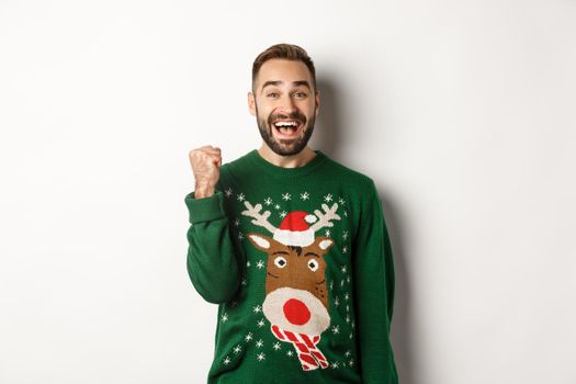 New Year, holidays and celebration. Excited and happy guy in Christmas sweater making fist pump, triumphing, receiving a gift and rejoicing, standing over white background.