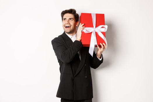 Concept of christmas holidays, celebration and lifestyle. Image of excited man enjoying new year, shaking gift box to guess what inside, standing against white background.