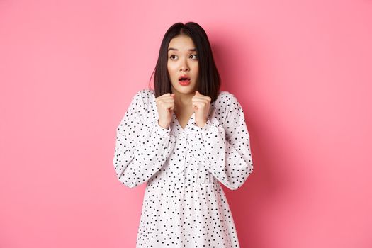 Scared and timid asian girl trembling from fear, staring left and gasping, standing in dress over pink background.