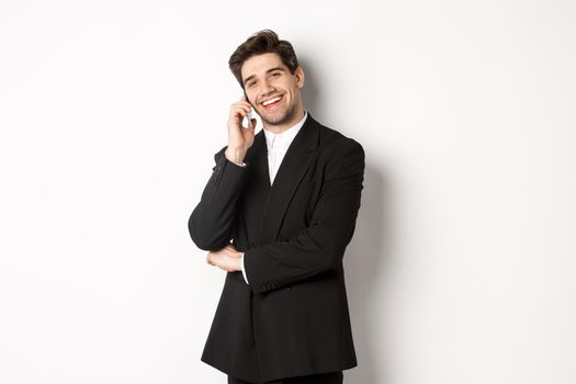 Image of handsome, successful businessman talking on phone, smiling pleased, standing in suit against white background.