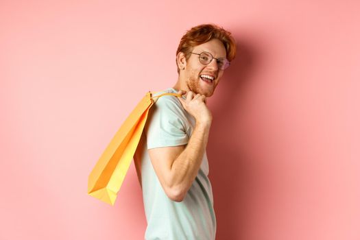 Carefree young man with red hair and glasses, walking with shopping bag over his shoulder and smiling, shopper buying presents, standing satisfied over pink background.