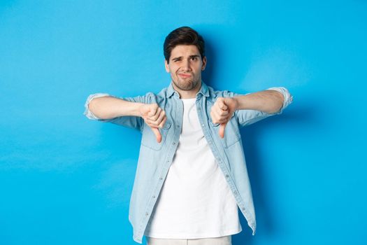 Skeptical and disappointed 25 years old man in casual outfit dislike something bad, showing thumbs-down and grimacing displeased, standing against blue background.