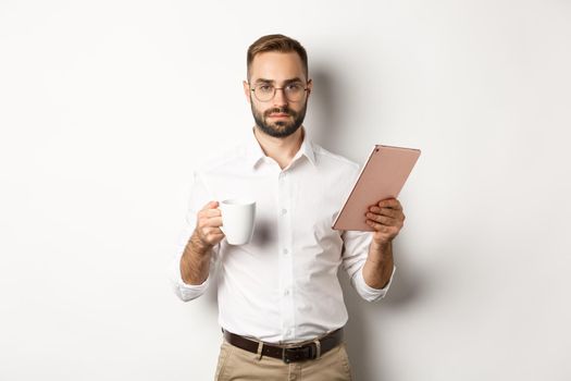 Confident male manager reading work on digital tablet and drinking coffee, standing over white background.