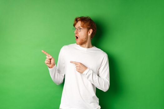 Impressed redhead man in glasses checking out promo, pointing and looking left with amazed face, standing in long-sleeve t-shirt over green background.