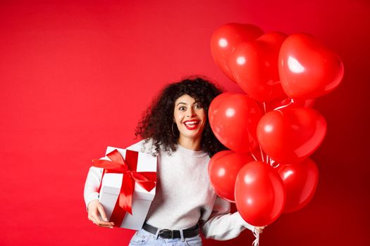 Holidays and celebration. Happy birthday girl holding gift and posing near party helium balloons, smiling excited at camera, red background.