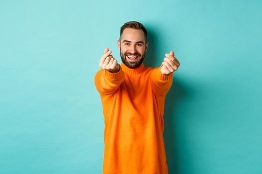 Smiling handsome man showing hearts and looking happy, lucky gesture, standing in orange sweater over light blue background.