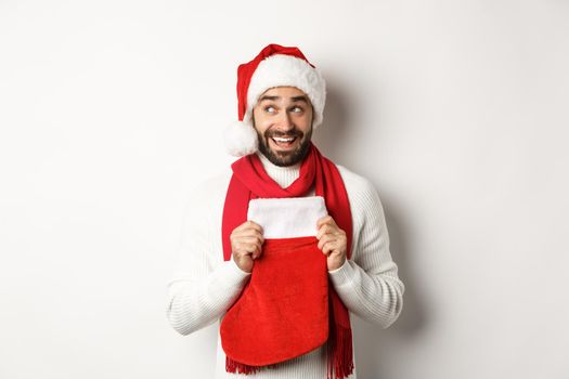 Winter holidays and shopping concept. Excited handsome man planning new year, holding Christmas sock for gifts and thinking, standing over white background.