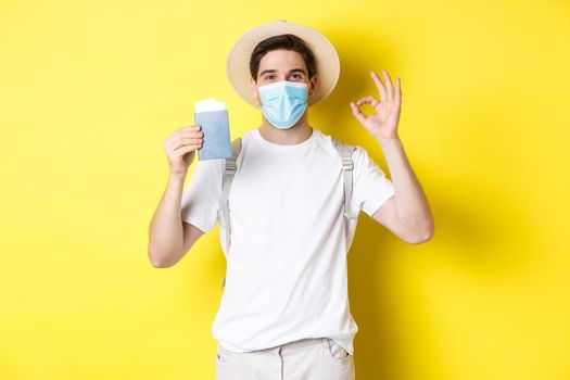 Concept of covid-19, tourism and pandemic. Happy male tourist in medical mask showing passport, going on vacation during coronavirus, make ok sign, yellow background.