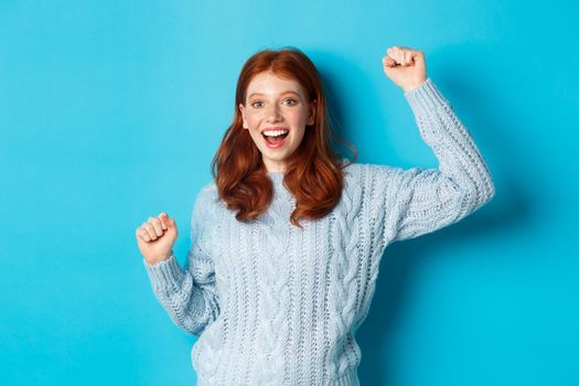 Cheerful redhead gil winning, celebrating victory, smiling and jumping from happiness, posing against blue background.