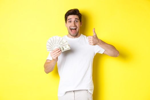 Excited man holding money, showing thumb-up in approval, got credit or loan, standing over yellow background.