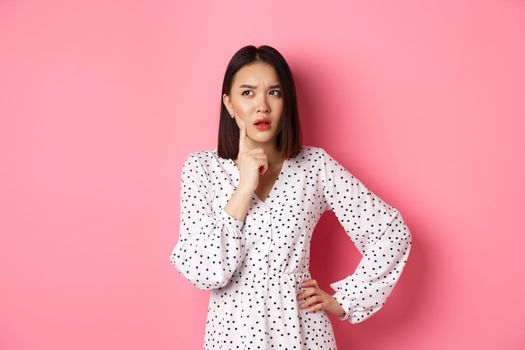 Thoughtful asian woman making assumption, looking up and thinking, deciding something, standing in dress over pink background.
