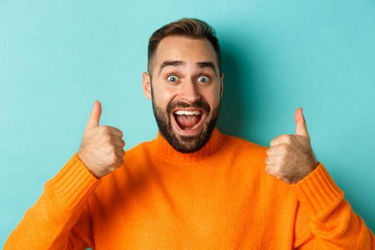 Close-up of excited handsome man showing thumb-up, recommending product, praising great quality, standing over light blue background.