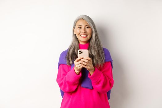 Happy asian woman smiling at camera after reading promotion on smartphone, standing with mobile phone over white background.