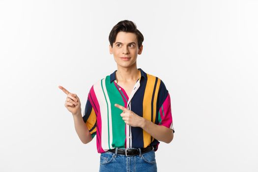 Young handsome queer man pointing, looking left at logo, smiling pleased, standing over white background.