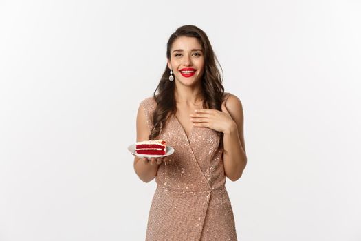 Party and celebration concept. Attractive slim woman in elegant dress holding piece of cake and smiling, standing over white background.