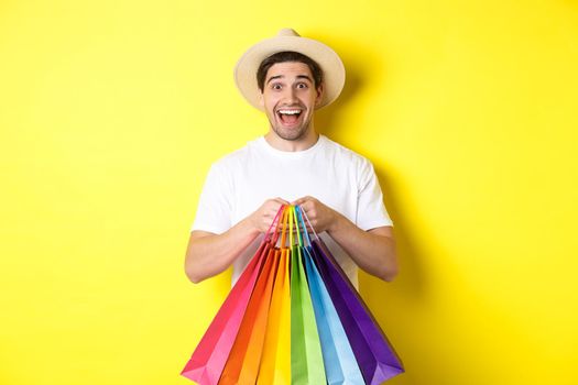 Image of happy man shopping on vacation, holding paper bags and smiling, standing against yellow background.