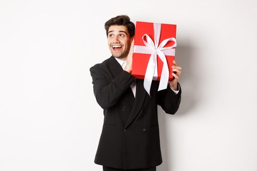 Image of handsome dreamy guy in black suit, shaking box with present to wonder whats inside, standing against white background happy.