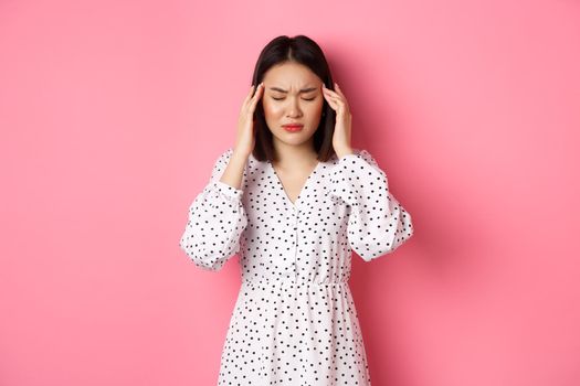 Image of upset asian woman having headache, feeling unwell or dizzy, close eyes and massaging head, suffering migraine, standing over pink background.