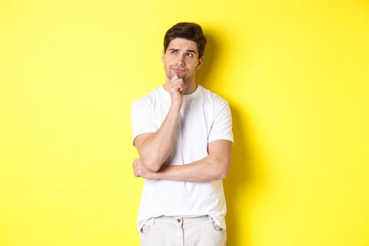 Portrait of young male model thinking, looking at upper left corner and making choice, standing near copy space, yellow background.