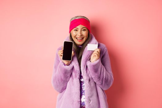Online shopping and fashion concept. Beautiful asian mature woman showing plastic credit card and blank phone screen, smiling happy, pink background.