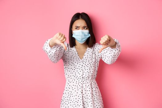 Coronavirus, social distancing and lifestyle concept. Disappointed asian woman in face mask looking judgemental, showing thumbs-down, express negative opinion, pink background.