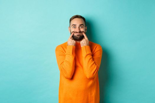 Image of bearded man stretching lips in happy smile, faking happiness, standing over light blue background.