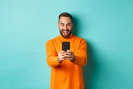 Image of man reading awesome message on mobile phone, looking at smartphone screen with amazement, standing over light blue background.