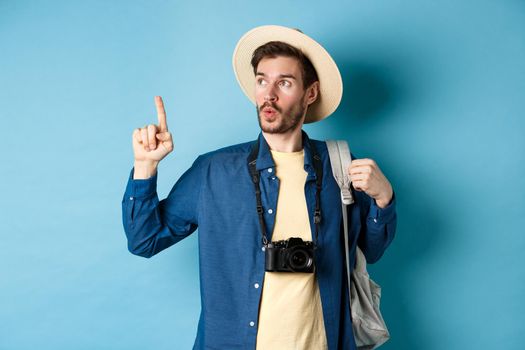 Handsome tourist looking curious and pointing up, showing interesting sightseeing on summer vacation, wearing straw hat and holding backpack with camera, blue background.
